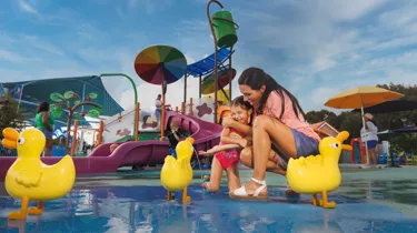 A mom and daughter enjoy a fun moment together at the Muddy Puddles Splash Pad at Peppa Pig Theme Park in Dallas Fort Worth. The daughter splashes in the water while the mom watches, both smiling and laughing in the playful, water-filled attraction.