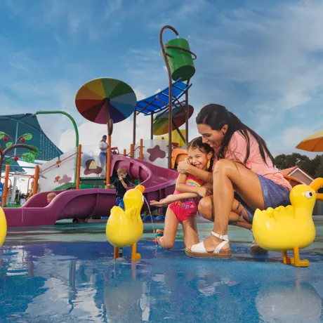 A mom and daughter enjoy a fun moment together at the Muddy Puddles Splash Pad at Peppa Pig Theme Park in Dallas Fort Worth. The daughter splashes in the water while the mom watches, both smiling and laughing in the playful, water-filled attraction.