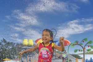 A little girl splashes around at the Muddy Puddles Splash Pad at Peppa Pig Theme Park in Dallas Fort Worth. She smiles and enjoys the playful water features, surrounded by the colorful, fun atmosphere of the park.