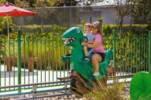 A mother and son enjoy riding Grampy Rabbit's Dinosaur Adventure at Peppa Pig Theme Park in Dallas Fort Worth. They smile and laugh as they embark on the exciting, prehistoric-themed ride, surrounded by the colorful and fun atmosphere of the park.