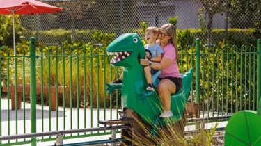 A mother and son enjoy riding Grampy Rabbit's Dinosaur Adventure at Peppa Pig Theme Park in Dallas Fort Worth. They smile and laugh as they embark on the exciting, prehistoric-themed ride, surrounded by the colorful and fun atmosphere of the park.