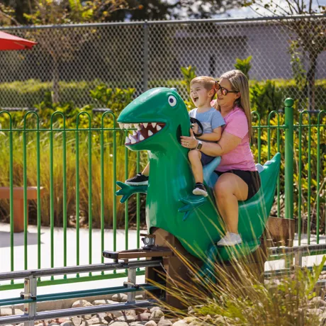 A mother and son enjoy riding Grampy Rabbit's Dinosaur Adventure at Peppa Pig Theme Park in Dallas Fort Worth. They smile and laugh as they embark on the exciting, prehistoric-themed ride, surrounded by the colorful and fun atmosphere of the park.