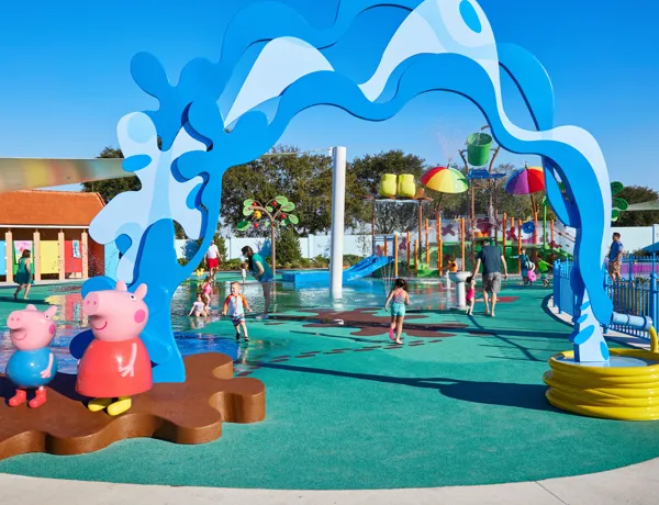 Niños y padres disfrutan del área de chapoteo Muddy Puddles en Peppa Pig Theme Park en Dallas Fort Worth. Los niños salpican el agua mientras los padres observan y se ríen, todo rodeado por la atmósfera juguetona y llena de agua del parque.