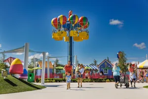 Un grupo de familias disfruta de la atracción Fun Fair en Peppa Pig Theme Park en Dallas Fort Worth, jugando y subiendo a diversas atracciones. Al fondo, se puede ver el paseo en Globo de Peppa, que añade a la vibrante y juguetona atmósfera del parque.